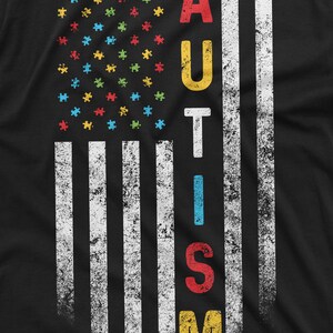 Peut inclure: T-shirt noir avec un motif drapeau am&eacute;ricain vieilli. Les &eacute;toiles sont remplac&eacute;es par des pi&egrave;ces de puzzle color&eacute;es et le mot "AUTISM" est imprim&eacute; verticalement en lettres color&eacute;es et en gras.