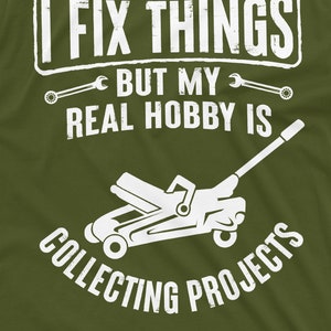 May include: A green t-shirt with white text that reads "I fix things but my real hobby is collecting projects". A white silhouette of a car jack is below the text.