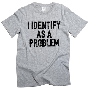 Puede incluir: Camiseta gris jaspeado con la frase "I IDENTIFY AS A PROBLEM" impresa en negro. La camiseta tiene mangas cortas y cuello redondo.