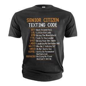 Peut inclure: Un t-shirt noir avec un graphique blanc qui dit "Senior Citizen Texting Code" et liste des abréviations de textos courantes utilisées par les personnes âgées. Les abréviations sont en texte orange.