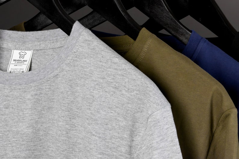 May include: Three t-shirts hanging on hangers. The t-shirts are gray, olive green, and navy blue. The gray t-shirt has a small white tag with the text "Silver Lake T-Shirts" and a graphic of a chef's hat.