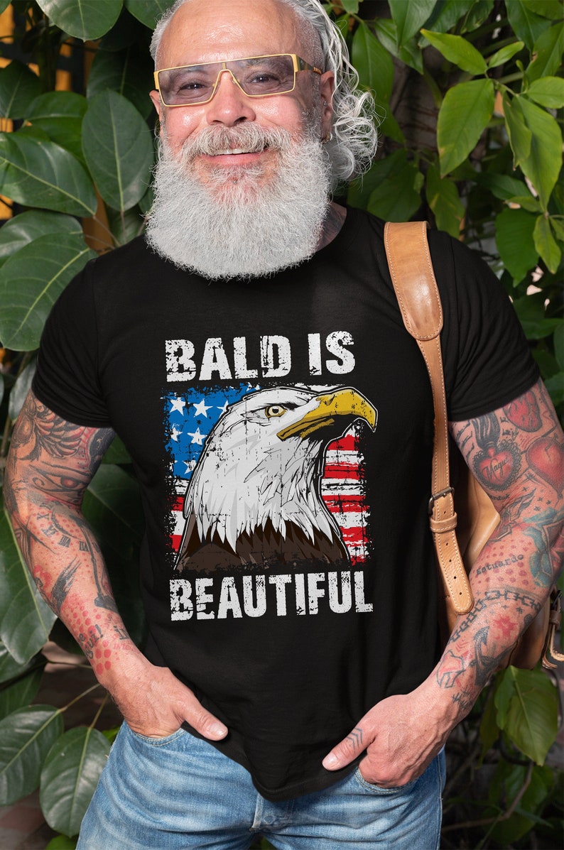 May include: A black t-shirt with a distressed American flag design and the text "Bald is Beautiful" in white lettering. An eagle with a distressed look is centered on the flag.