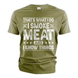 May include: A green t-shirt with a white graphic that says "That's what I do I smoke meat and I know things". The graphic also includes two crossed grilling forks and a flame.