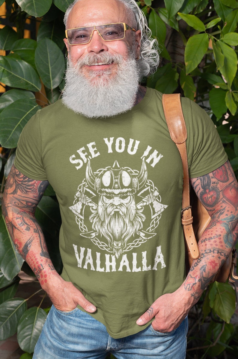 May include: Olive green t-shirt with a white graphic of a Viking warrior and the text "SEE YOU IN VALHALLA". The design includes a horned helmet, axes, and a beard. The shirt is worn by a person with tattoos.