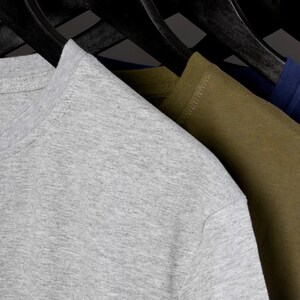 May include: Three t-shirts hanging on hangers. The t-shirts are gray, olive green, and navy blue. The gray t-shirt has a small white tag with the text "Silver Lake T-Shirts" and a graphic of a chef's hat.