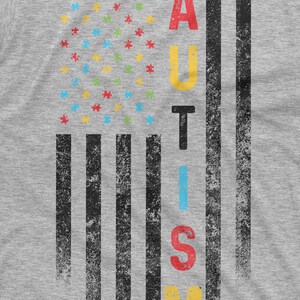 Peut inclure: Un t-shirt gris avec un motif vieilli. Le motif pr&eacute;sente un graphique en forme de drapeau avec des pi&egrave;ces de puzzle &agrave; la place des &eacute;toiles et le mot "AUTISME" en lettres color&eacute;es. Les rayures du drapeau sont noires.