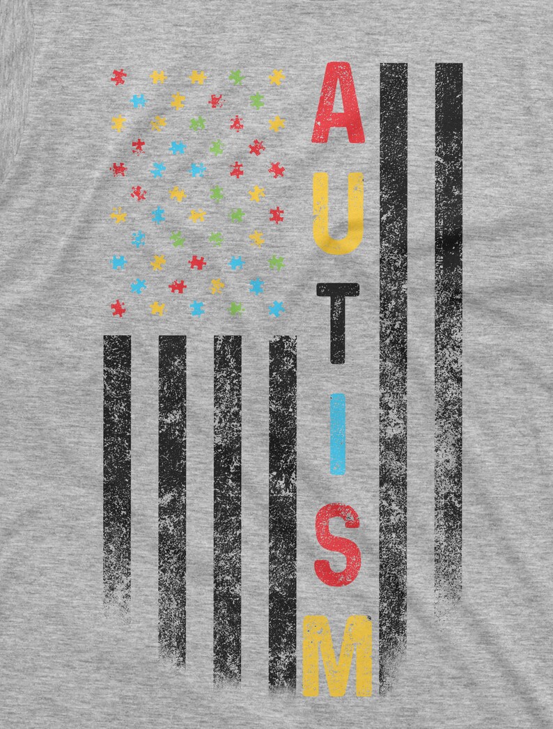 Peut inclure: Un t-shirt gris avec un motif vieilli. Le motif pr&eacute;sente un graphique en forme de drapeau avec des pi&egrave;ces de puzzle &agrave; la place des &eacute;toiles et le mot "AUTISME" en lettres color&eacute;es. Les rayures du drapeau sont noires.