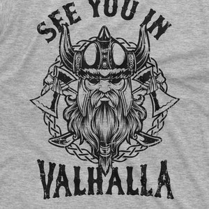 May include: A gray t-shirt featuring a black and white illustration of a Viking warrior with a horned helmet, beard, and crossed axes. The text "SEE YOU IN VALHALLA" is printed above and below the warrior's image.