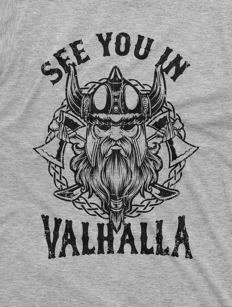 May include: A gray t-shirt featuring a black and white illustration of a Viking warrior with a horned helmet, beard, and crossed axes. The text "SEE YOU IN VALHALLA" is printed above and below the warrior's image.