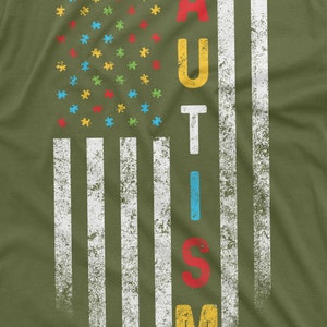Peut inclure: T-shirt vert olive avec un motif vieilli du drapeau am&eacute;ricain. Les &eacute;toiles sont remplac&eacute;es par des pi&egrave;ces de puzzle color&eacute;es, et le mot "AUTISM" est affich&eacute; verticalement. Le design a un aspect vintage et us&eacute;.