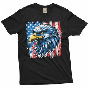 May include: Black t-shirt featuring a graphic of an American bald eagle with blue and white feathers and a yellow beak. The eagle is set against a backdrop of the American flag in red, white, and blue.
