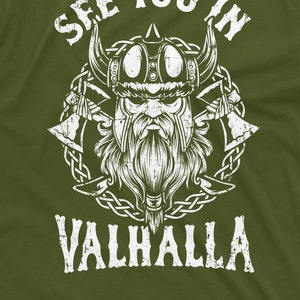 May include: Olive green t-shirt featuring a distressed white graphic of a Viking with a horned helmet, beard, and crossed axes. The text "SEE YOU IN VALHALLA" is arched above and below the Viking illustration.