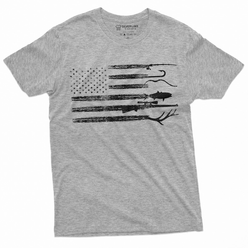 May include: A heather grey t-shirt with a distressed American flag design featuring fishing and hunting imagery. The flag is made up of black silhouettes of fishing rods, hooks, a fish, a rifle, and antlers.