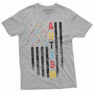 Peut inclure: Un t-shirt gris chin&eacute; avec un motif vieilli. Le motif repr&eacute;sente un drapeau am&eacute;ricain stylis&eacute; avec des pi&egrave;ces de puzzle &agrave; la place des &eacute;toiles et le mot "AUTISM" en lettres color&eacute;es. Le t-shirt est de Silver Lake T-shirts.