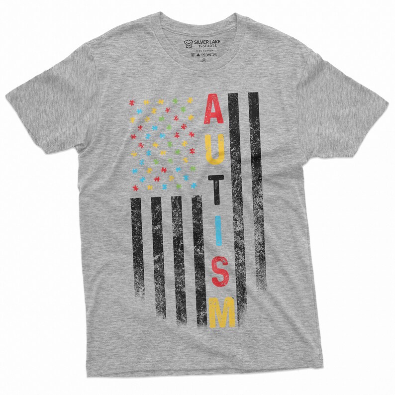 Peut inclure: Un t-shirt gris chin&eacute; avec un motif vieilli. Le motif repr&eacute;sente un drapeau am&eacute;ricain stylis&eacute; avec des pi&egrave;ces de puzzle &agrave; la place des &eacute;toiles et le mot "AUTISM" en lettres color&eacute;es. Le t-shirt est de Silver Lake T-shirts.