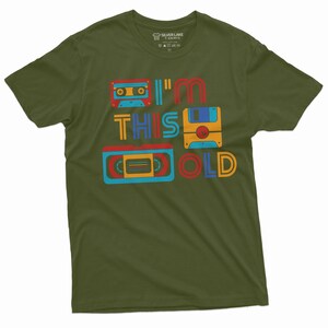 May include: A green t-shirt with a retro design featuring a cassette tape, floppy disk, and VHS tape. The text on the shirt reads "I'm This Old".