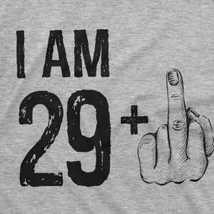 May include: A gray t-shirt with the text "I AM 29+" printed on it. The number 29 is in a large, bold font and the plus sign is next to it. A hand with the middle finger extended is printed on the right side of the shirt.