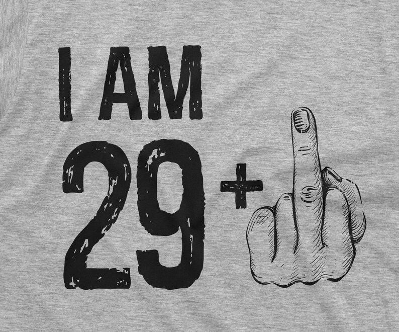 May include: A gray t-shirt with the text "I AM 29+" printed on it. The number 29 is in a large, bold font and the plus sign is next to it. A hand with the middle finger extended is printed on the right side of the shirt.