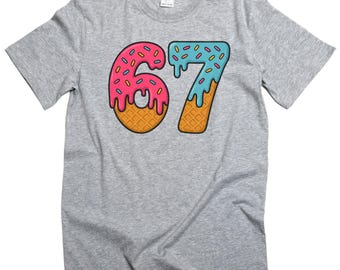 May include: A heather gray t-shirt featuring a colorful graphic of the number "67". The numbers are designed to look like ice cream cones with pink and blue frosting, sprinkles, and waffle cone bases. The shirt is made of a soft material.