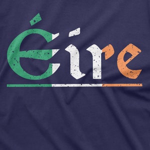 May include: A navy blue t-shirt with a distressed white and orange graphic of the word "&Eacute;ire" in a traditional Irish font. The graphic is layered with a green, white, and orange stripe, resembling the Irish flag.
