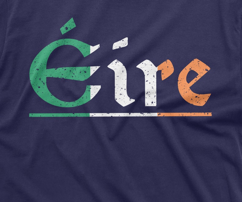May include: A navy blue t-shirt with a distressed white and orange graphic of the word "&Eacute;ire" in a traditional Irish font. The graphic is layered with a green, white, and orange stripe, resembling the Irish flag.
