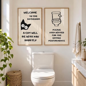Könnte beinhalten: Zwei rechteckige Wandbilder in hellen Holzrahmen mit cremefarbenem Hintergrund und schwarzem Text. Das linke Bild zeigt eine Katzenillustration und den Text: WELCOME TO THE BATHROOM A CAT WILL BE WITH YOU SHORTLY. Das rechte Bild zeigt ein Toilettensymbol und den Text: PLEASE STAY SEATED FOR THE ENTIRE PERFORMANCE.
