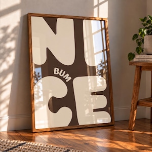 Könnte beinhalten: Ein rechteckiges gerahmtes Wandbild mit dunkelbraunem Hintergrund und großen, fetten, cremefarbenen Buchstaben, die das Wort NICE bilden, wobei das Wort BUM in kleineren cremefarbenen Buchstaben zwischen dem N und dem C steht.