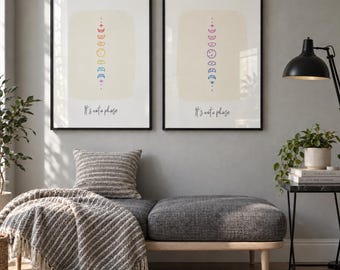 Op de afbeelding: Twee ingelijste wandposters met zwarte lijsten, elk met een verticale reeks maanfasen in een regenboogverloop op een lichtbeige achtergrond, met de tekst "It's not a phase" in zwarte schrijfletters onder het ontwerp.
