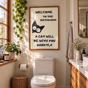 Könnte beinhalten: Ein rechteckiges Wandbild in einem Holzrahmen mit einer schwarzen Katzengrafik, die von der linken Seite hervorlugt. Der Text lautet: WELCOME TO THE BATHROOM A CAT WILL BE WITH YOU SHORTLY. Der Hintergrund ist cremefarben mit schwarzer Schrift.