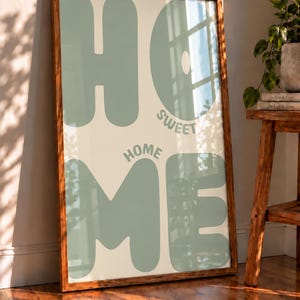 Könnte beinhalten: Ein rechteckiger Kunstdruck in einem dunklen Holzrahmen mit dem Wort HOME in großen, salbeigrünen, abgerundeten Buchstaben. Die Wörter SWEET HOME sind in kleinerer, salbeigrüner Schrift zwischen den großen Buchstaben auf cremefarbenem Grund.