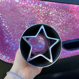 May include: A silver star-shaped object inside a black cylinder. The star is centered within the cylinder. The background is a pink surface with a pattern of silver stars.