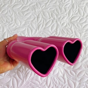 May include: A pair of pink ceramic vases shaped like hearts with black interiors. The vases are glossy and have a smooth finish.