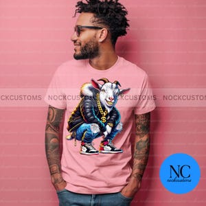 May include: A pink t-shirt with a cartoon image of a goat wearing a black leather jacket, blue jeans, and gold chains. The goat is sitting with its legs crossed and has a gold dollar sign on its chest.