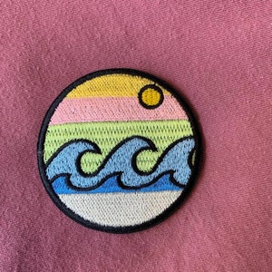 May include: A round embroidered patch with a yellow sun, pink, green, and blue stripes, and blue waves. The patch is on a pink fabric background.