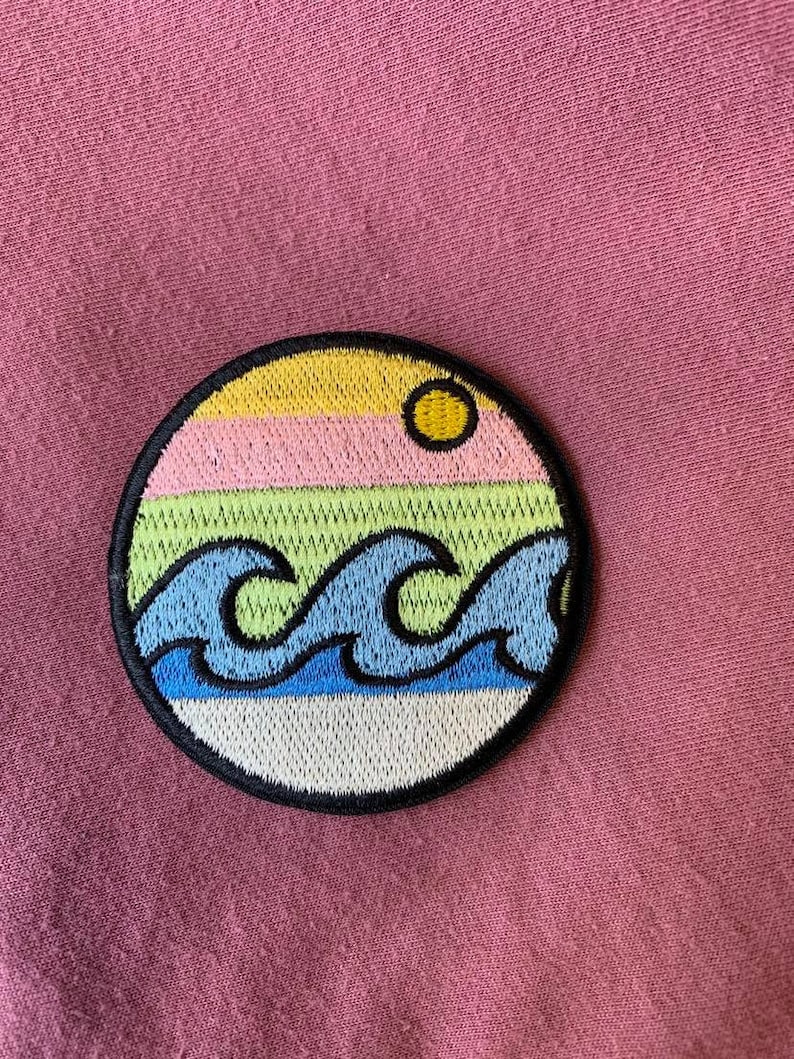 May include: A round embroidered patch with a yellow sun, pink, green, and blue stripes, and blue waves. The patch is on a pink fabric background.