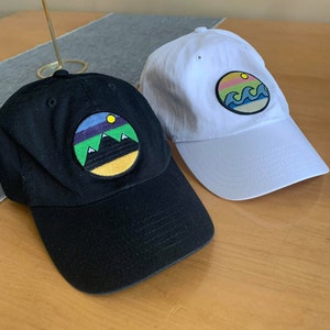 May include: Two baseball caps with embroidered patches. One cap is black with a mountain scene patch, the other is white with a wave scene patch.