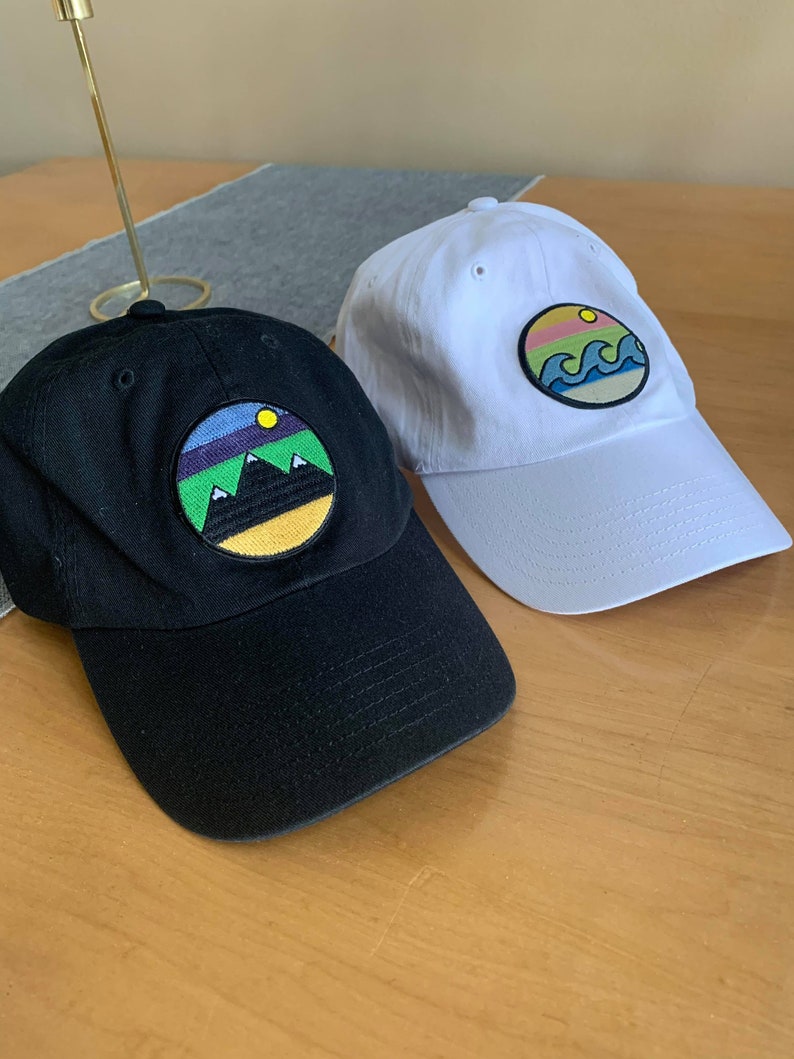 May include: Two baseball caps with embroidered patches. One cap is black with a mountain scene patch, the other is white with a wave scene patch.