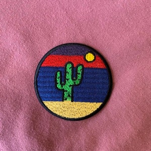May include: A round embroidered patch with a sunset scene and a green cactus in the foreground. The sunset is depicted with red, orange, yellow, blue, and purple colors. The patch has a black outline.