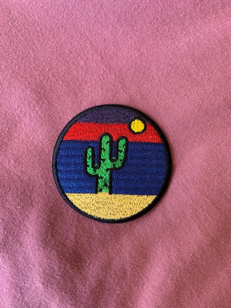 May include: A round embroidered patch with a sunset scene and a green cactus in the foreground. The sunset is depicted with red, orange, yellow, blue, and purple colors. The patch has a black outline.
