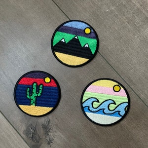 May include: Three embroidered iron-on patches featuring different nature scenes. The first patch depicts a yellow, green, and blue mountain scene with a small yellow sun. The second patch shows a yellow, red, and blue desert scene with a green cactus. The third patch is a yellow, pink, green, and blue ocean scene with blue waves.