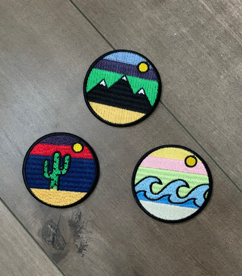 May include: Three embroidered iron-on patches with different nature scenes. The first patch is a yellow, green, and blue mountain scene with a small yellow sun. The second patch is a yellow, red, and blue desert scene with a green cactus. The third patch is a yellow, pink, green, and blue ocean scene with blue waves.