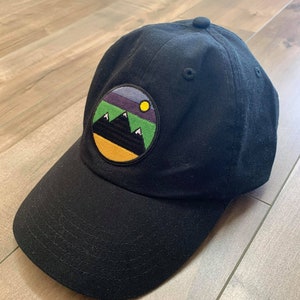 May include: A black baseball cap with a round, colorful patch featuring a mountain range and a yellow sun. The patch has a green, purple, and blue gradient.