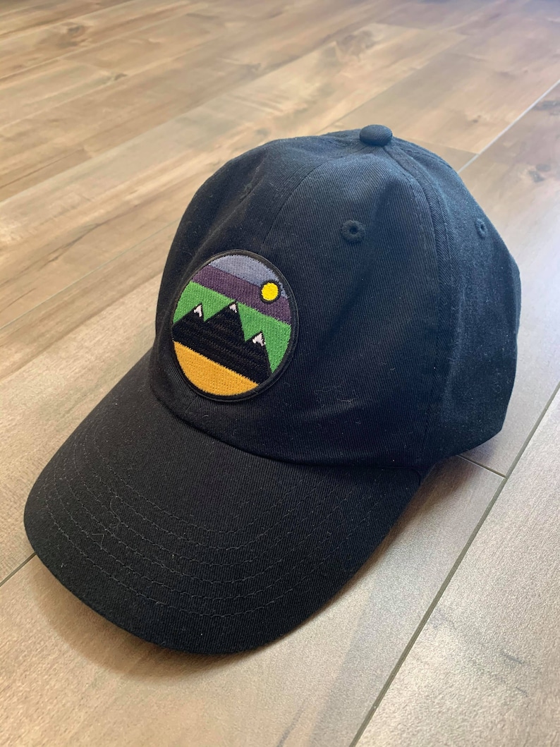 May include: A black baseball cap with a round, colorful patch featuring a mountain range and a yellow sun. The patch has a green, purple, and blue gradient.