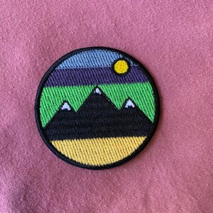 May include: A round embroidered patch with a black mountain range, green grass, a yellow sun, and a blue and purple sky.