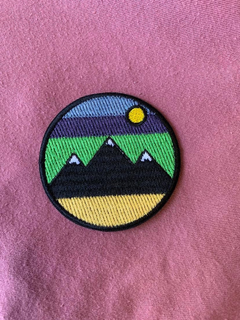 May include: A round embroidered patch with a black mountain range, green grass, a yellow sun, and a blue and purple sky.