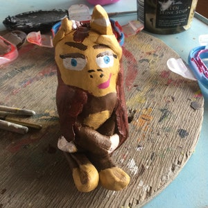 May include: A hand-painted clay figurine of a cartoon character with brown hair, a yellow face, blue eyes, and a pink mouth. The figurine is sitting with its legs crossed and its arms folded. The figurine is painted on a wooden surface with paint splatters.