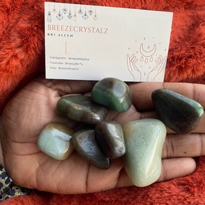 Puede incluir: Una colecci&oacute;n de piedras preciosas pulidas verdes y marrones de varias formas y tama&ntilde;os. Tambi&eacute;n se ve una tarjeta de visita con el texto "Breezecrystalz" y las redes sociales. Las piedras preciosas se muestran sobre una superficie roja y difusa.