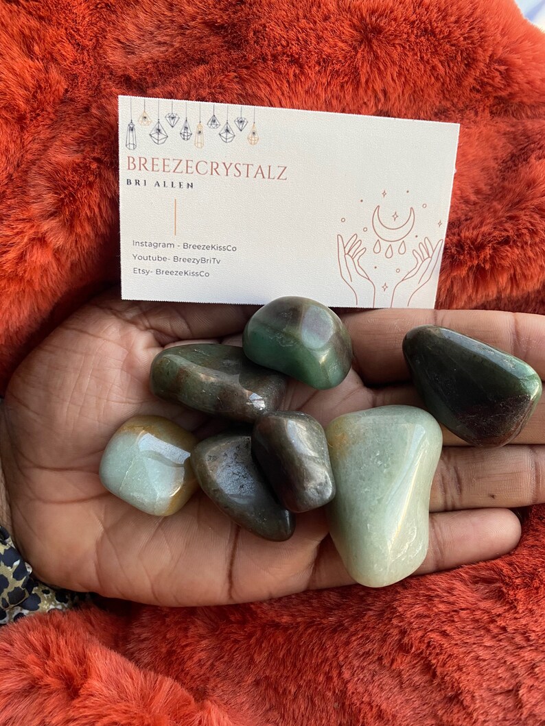 Puede incluir: Una colecci&oacute;n de piedras preciosas pulidas verdes y marrones de varias formas y tama&ntilde;os. Tambi&eacute;n se ve una tarjeta de visita con el texto "Breezecrystalz" y las redes sociales. Las piedras preciosas se muestran sobre una superficie roja y difusa.