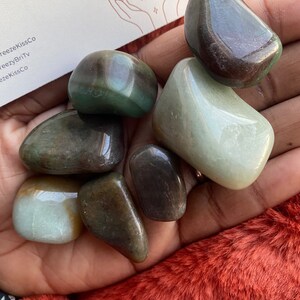 Puede incluir: Una colecci&oacute;n de piedras preciosas pulidas en tonos verdes, marrones y crema. Las piedras son lisas y redondeadas, con un acabado brillante. Una tarjeta de visita con el texto "Crystalz" es visible en el fondo.