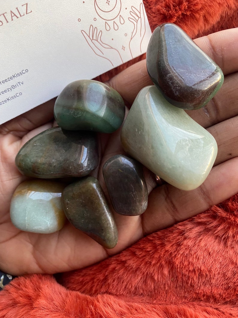 Puede incluir: Una colecci&oacute;n de piedras preciosas pulidas en tonos verdes, marrones y crema. Las piedras son lisas y redondeadas, con un acabado brillante. Una tarjeta de visita con el texto "Crystalz" es visible en el fondo.